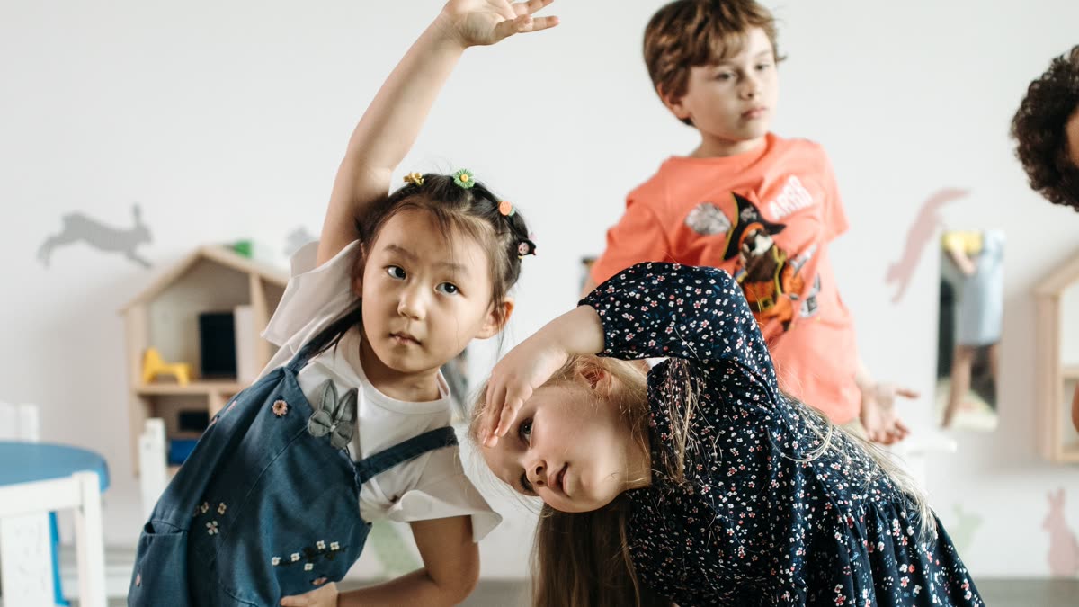 Three young children stretching and bending together in a playroom, mid-movement.