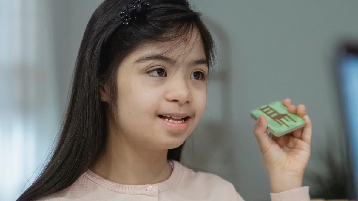 A girl at a desk holds up a picture tile mid-sentence, looking off to one side with rows of tiles laid out in front of her.