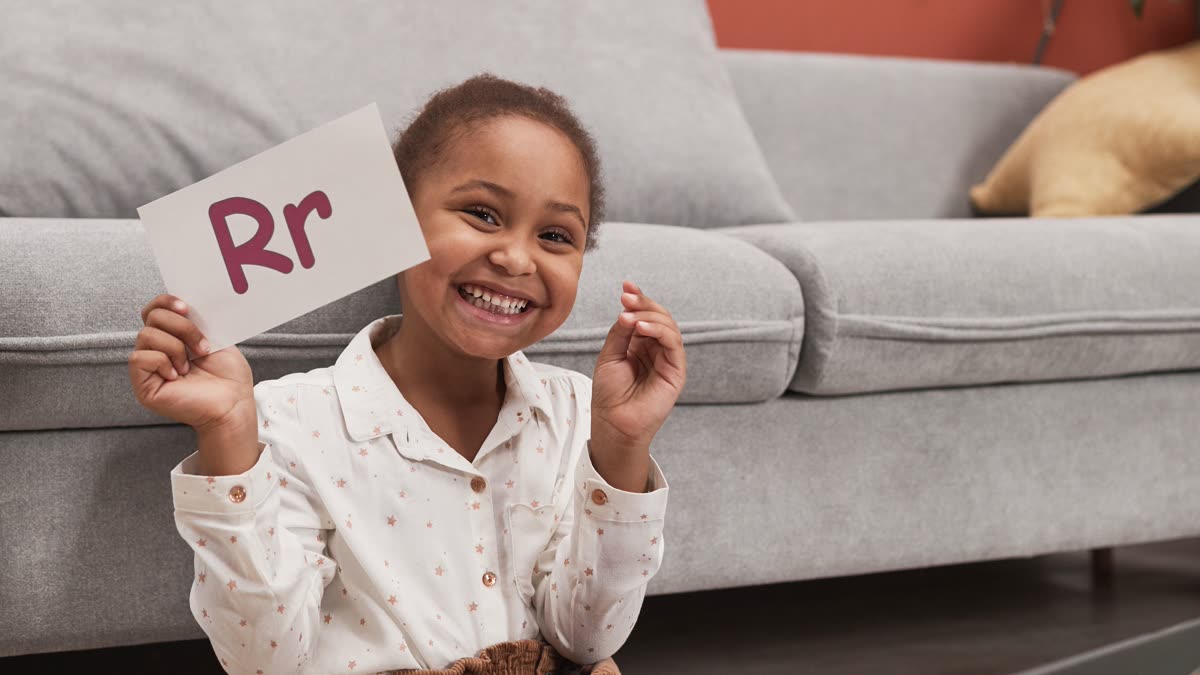 A smiling girl sits on the floor holding up a card printed with the letter R.