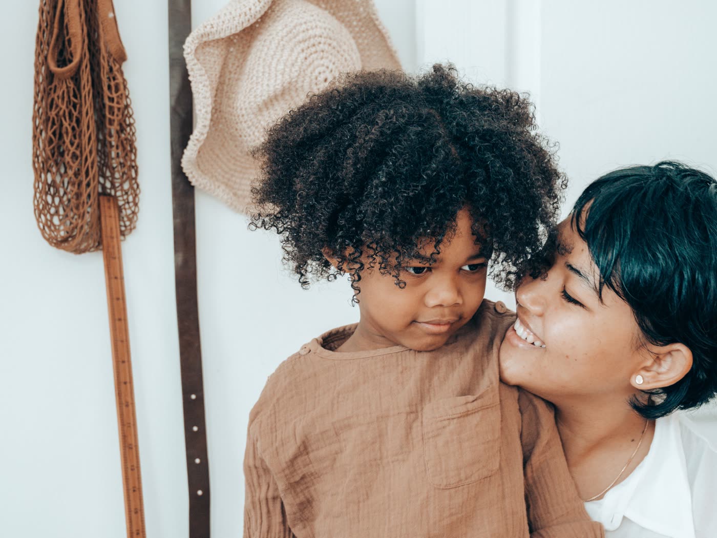 A young child leans back against an adult who holds them and smiles, in a hallway with a coat rack behind them.
