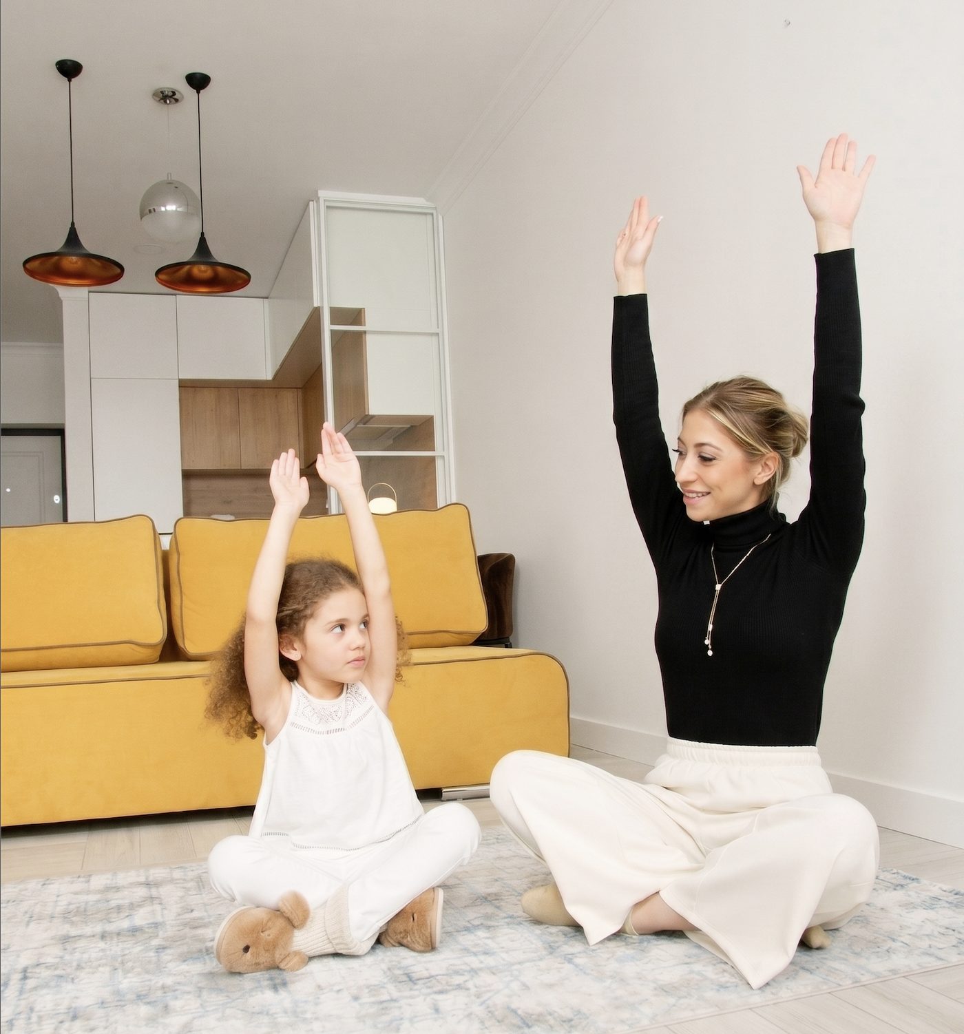 A young child and an adult sit cross-legged on the floor at home, both stretching their arms high overhead together.