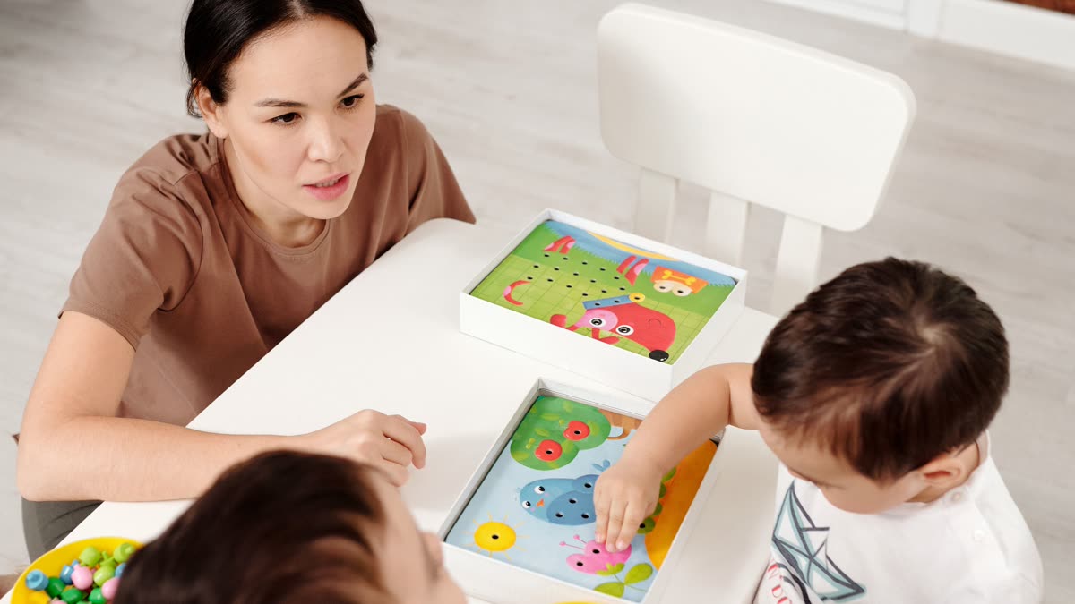 An adult sits at a low table with two young children, working through a picture-and-peg activity together.